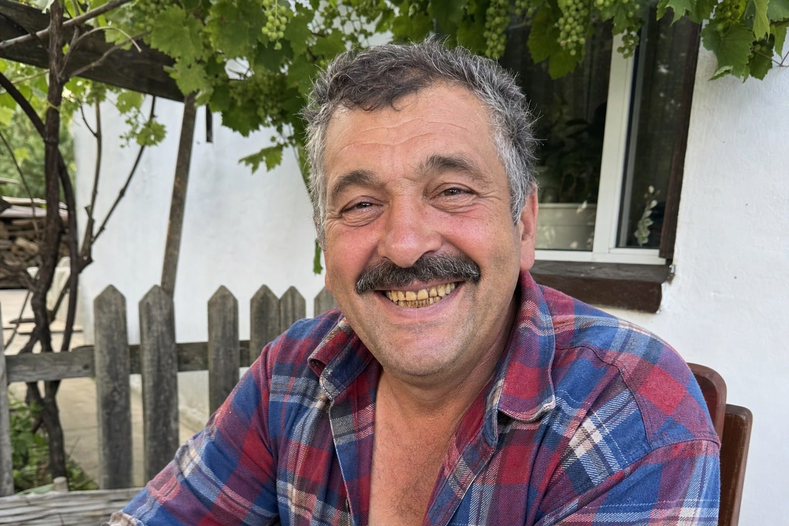 Andrei L., 41 ani, București — înainte de rutina Bright White, în curte la țară (clădire albă, gard, viță de vie), cămașă în carouri roșu-albastru, dinți galben-maronii de la tutun